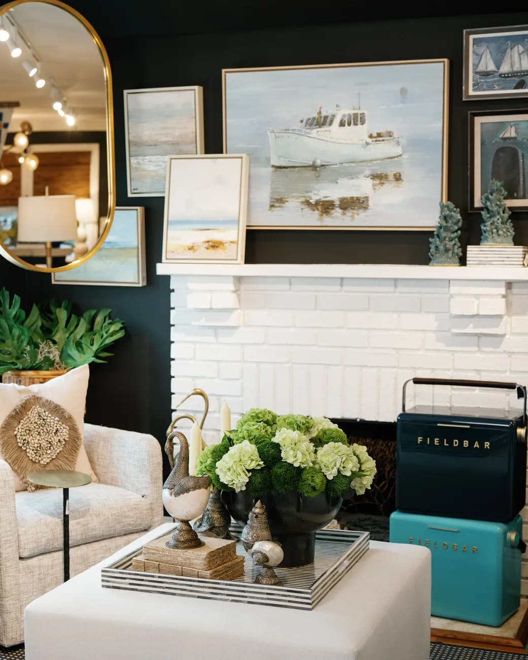 A styled bar vignette in the shop: framed harbor paintings on a white mantel, a turquoise Field Bar cooler chilling two bottles, green hydrangeas and a silver swan on the tray