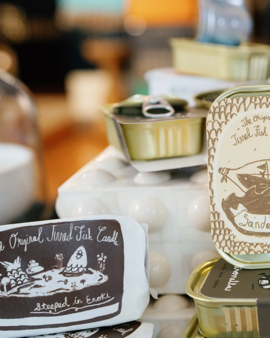 The Original Tinned Fish Candle display: gold sardine tins and hand-lettered Sandalwood labels stacked on a white milk-glass pedestal
