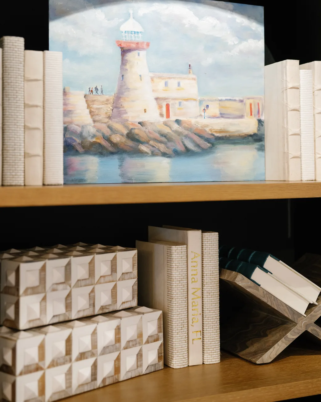 An oak shelf with a painted lighthouse harbor propped between white bookends, over a lower shelf of textured books lettered Anna Maria, FL