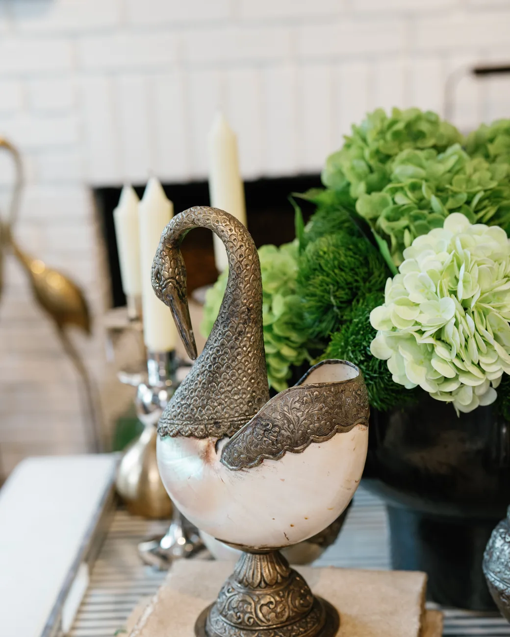 A tablescape detail: a silver-mounted nautilus swan vessel beside green hydrangeas and ivory taper candles on a white brick hearth