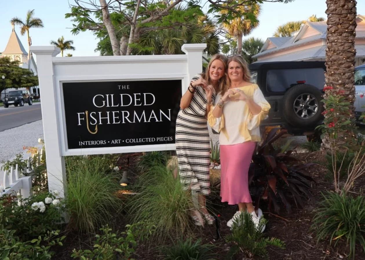 Two women celebrating beside the black Gilded Fisherman sign on Pine Avenue at dusk