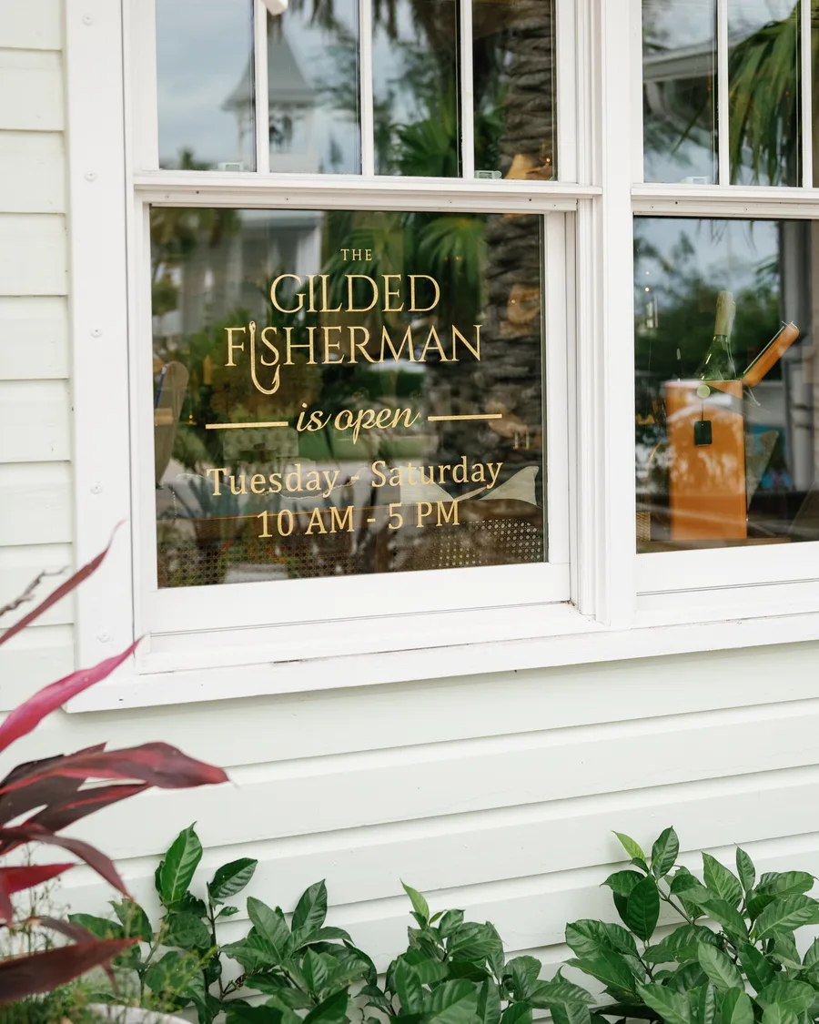 A front window in gilt lettering — The Gilded Fisherman is open Tuesday to Saturday, 10 AM to 5 PM — over a bed of red-leaved plants