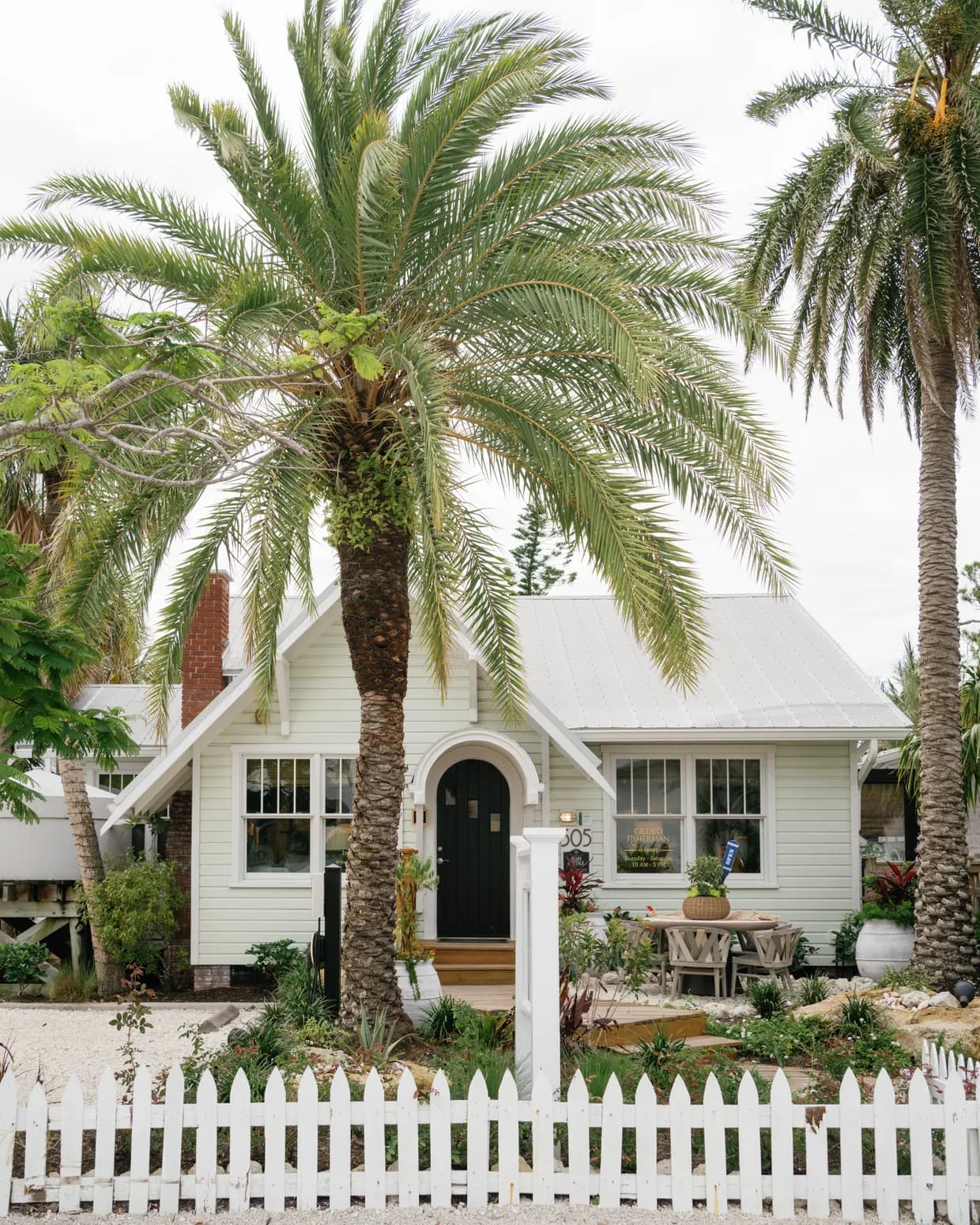 The Gilded Fisherman storefront: a pale-green island cottage with a black arched door and metal roof, framed by tall date palms behind a white picket fence